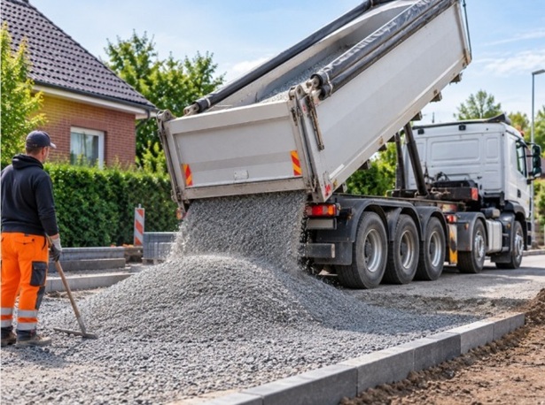 Lkw kippt feinen grauen Granitsplitt auf eine Baustelle für Pflasterarbeiten in Hamburg. Ein Mitarbeiter steht mit einem Rechen daneben.